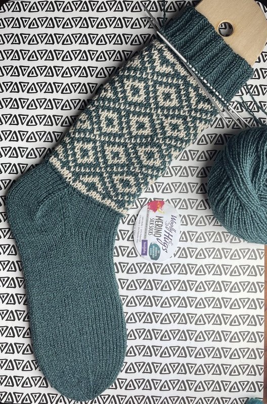 Gestrickte Socke in Petrol und Naturweiß mit Jacquardmuster auf Sockenspanner, Garnknäuel daneben