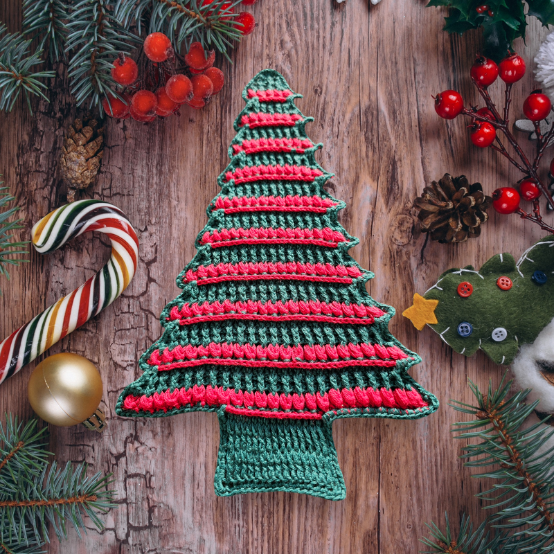 Nahaufnahme eines gehäkelten Tannenbaum-Deckchens mit roten Streifen auf Holzuntergrund und Weihnachtsakzenten