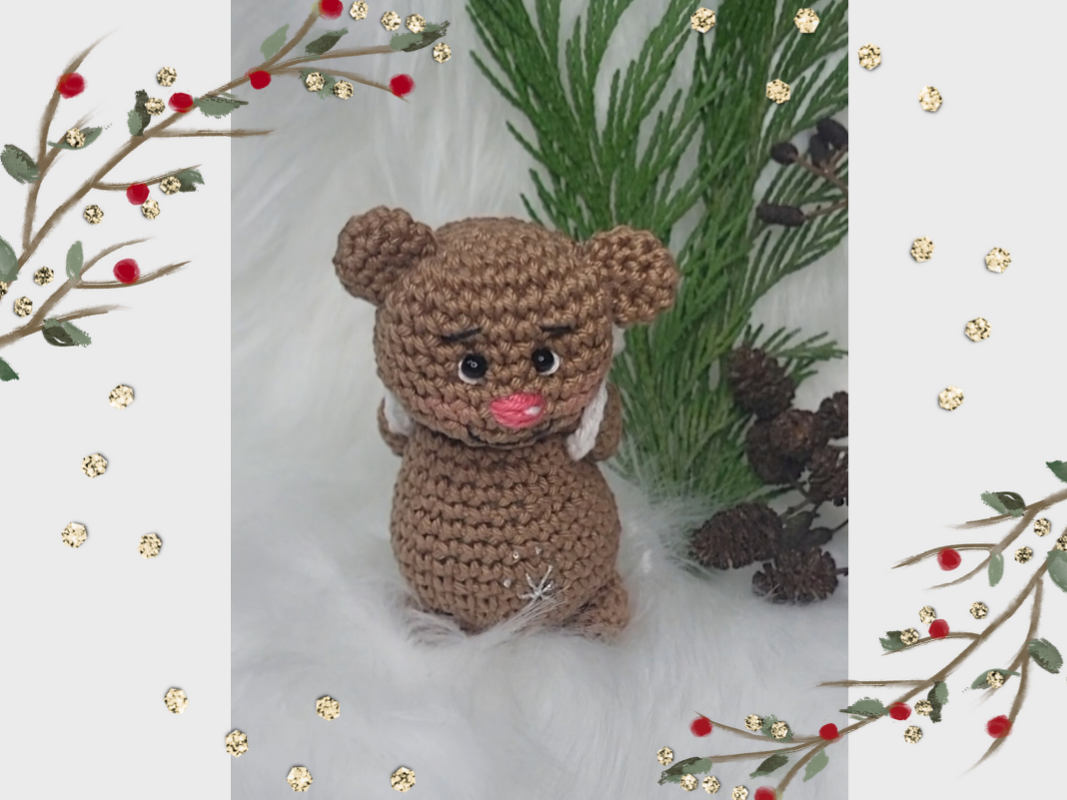 Kleiner brauner Amigurumi-Teddybär mit Schneeflocke und rosa Schnauze, umgeben von Tannenzweigen und Zapfen.