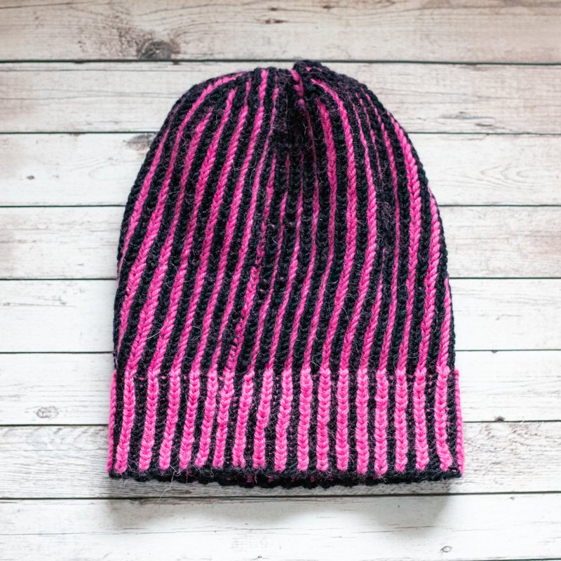 Zweifarbige gestrickte Damenmütze in Schwarz und Pink mit senkrechten Rippen auf hellem Holzuntergrund