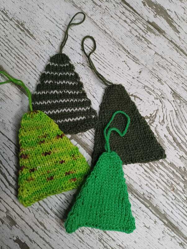 Vier gestrickte dreieckige Anhänger in verschiedenen Grüntönen mit Schlaufen auf hellem Holzuntergrund