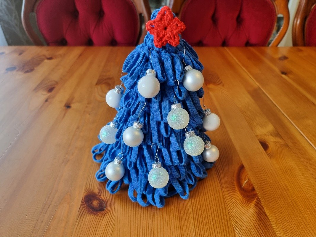 Blauer gehäkelter Mini-Tannenbaum mit weißen Kugeln und rotem Stern auf Holztisch