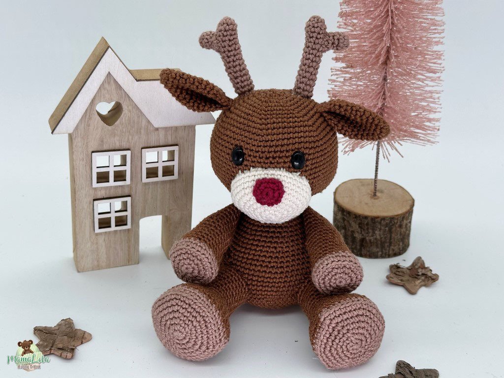 Braunes gehäkeltes Amigurumi-Rentier mit roter Nase vor Holzhaus und rosafarbenem Deko-Baum.