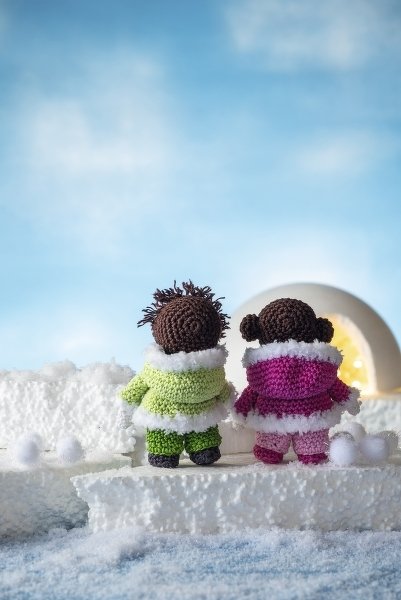 Zwei gehäkelte Amigurumi-Puppen mit grünen und pinken Wintermänteln vor einem beleuchteten Iglu.