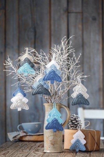 Gehäkelte Tannenbaum-Anhänger in Blau-, Grau- und Weißtönen an Zweigen in einer Vase neben Zapfen und Geschenk