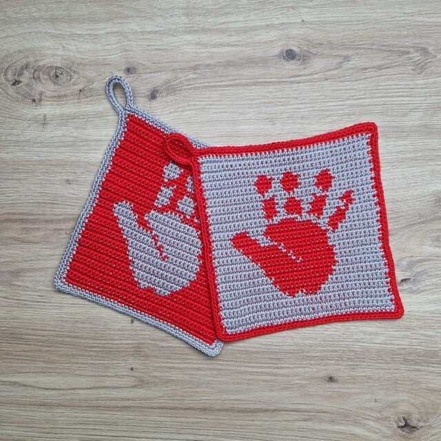 Zwei gehäkelte Topflappen mit kontrastierendem Handabdruck-Motiv in Rot und Grau.