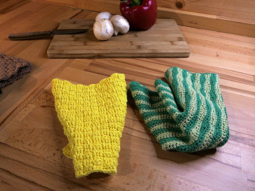 Gelber gehäkelter Topflappen und grün-gestreifter Ofenhandschuh auf einer Holzplatte, Schneidebrett mit Pilzen und Paprika im Hintergrund.