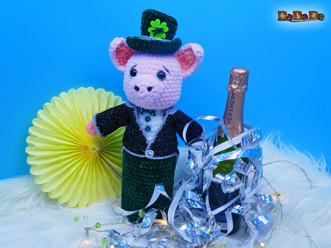Rosa gehäkeltes Schwein mit schwarzem Glitzeranzug und grünem Zylinder, daneben mit Lametta geschmückte Flasche auf weißem Fell vor blauem Hintergrund.