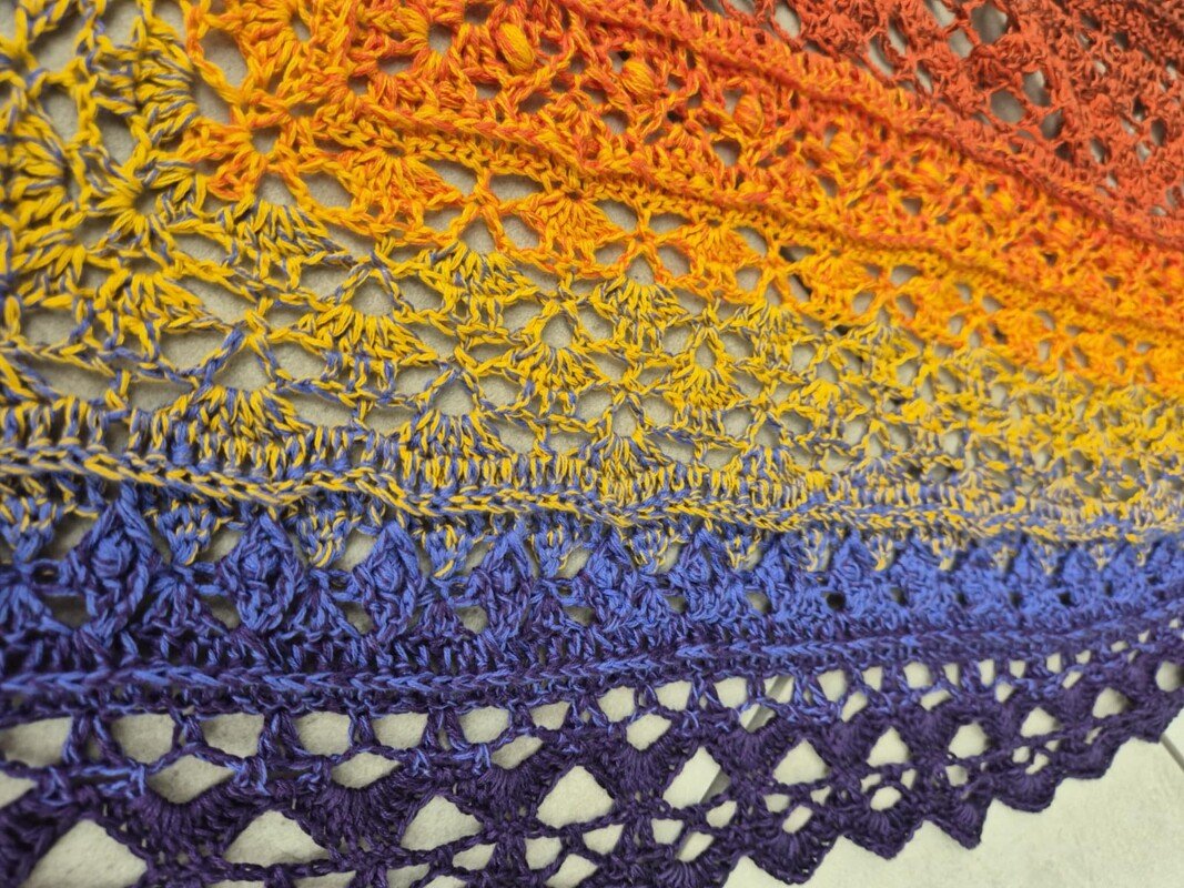 Crocheted triangle shawl with a gradient from orange to yellow to blue and purple, showing lacy openwork stitches and a scalloped edge.