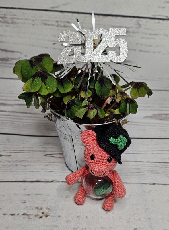 Rosa gehäkeltes Amigurumi‑Glücksschwein mit schwarzem Zylinderhut und grünem Kleeblatt vor einem Topf mit Glücksklee und einer 2025‑Dekoration.