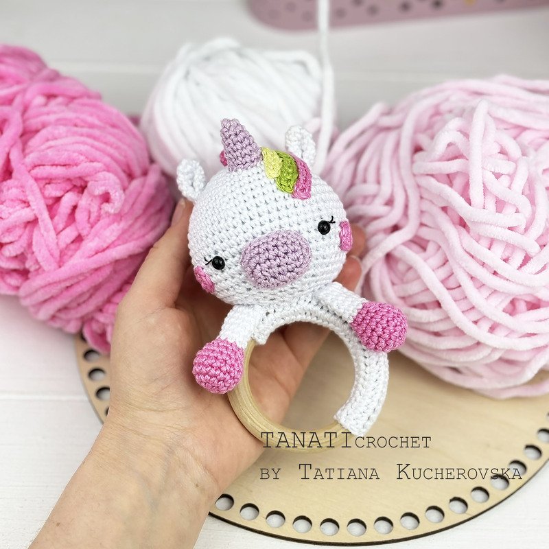 Nahaufnahme eines gehäkelten Einhorn-Greifrings in Weiß und Rosa mit Holzring, gehalten von einer Hand; rosa und weiße Garnknäuel im Hintergrund.