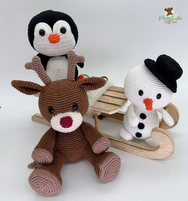 Gehäkelter Pinguin hinter einem gehäkelten Rentier und neben einem gehäkelten Schneemann mit schwarzem Hut, davor ein kleiner Holzschlitten