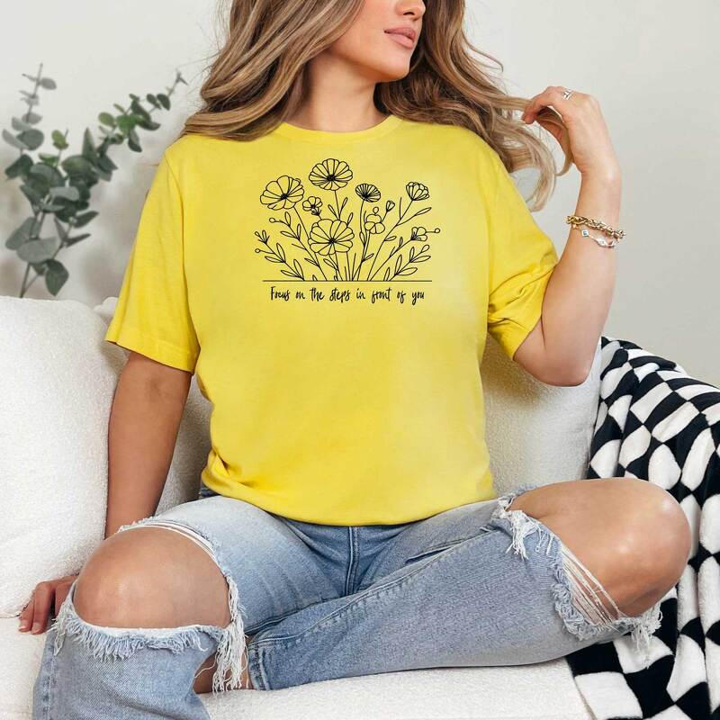 Gelbes T-Shirt mit schwarzer Blumen-Lineart und dem englischen Spruch „Focus on the steps in front of you“