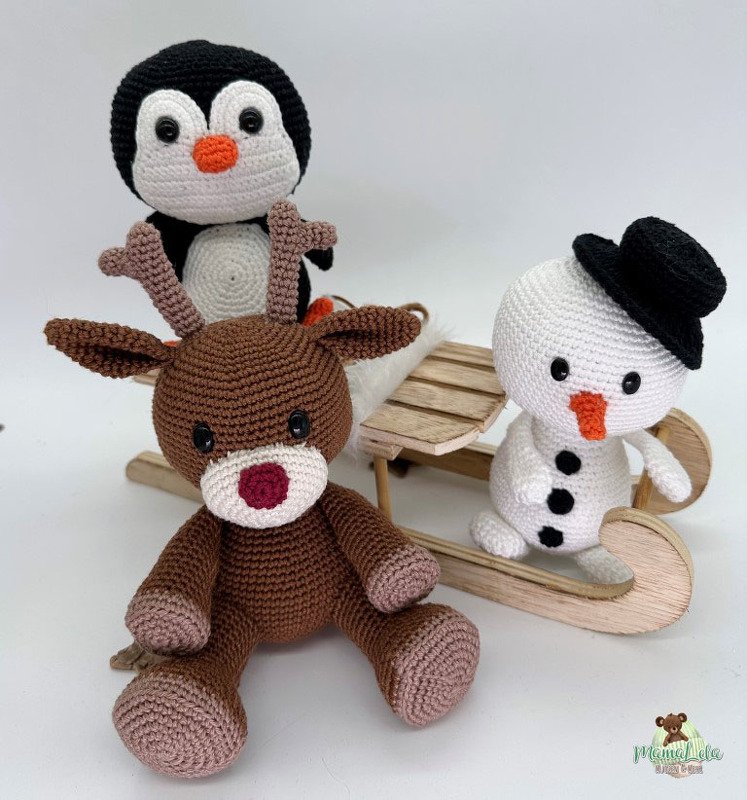 Gehäkeltes Rentier, Pinguin und Schneemann mit schwarzem Hut neben einem kleinen Holzschlitten