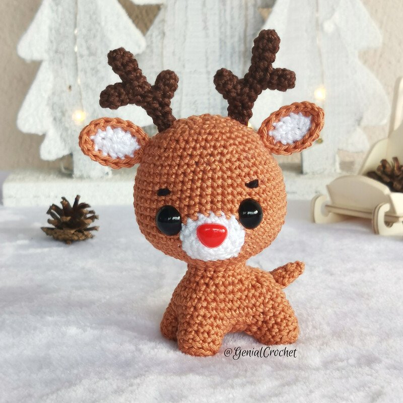 Small brown crochet reindeer amigurumi with a red nose and antlers on a white surface with winter decor.