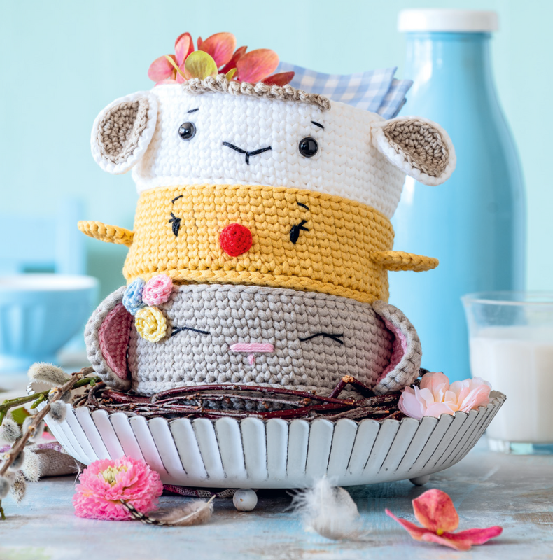 Drei gestapelte gehäkelte Tierkörbe im Amigurumi‑Stil mit Blumenapplikationen und Pastellfarben auf einer Schale