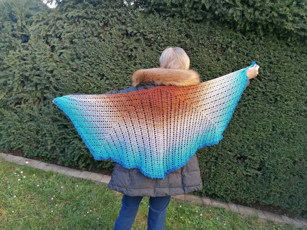 Rückansicht einer Person mit ausgestrecktem gehäkeltem Dreieckstuch in Blau, Weiß und Braun vor einer Hecke.