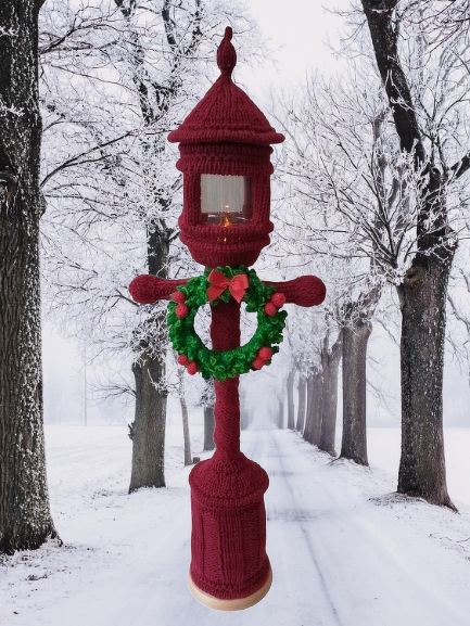 Maroon knitted lantern cover decorated with a green wreath and lit candle, set on a snowy tree-lined path