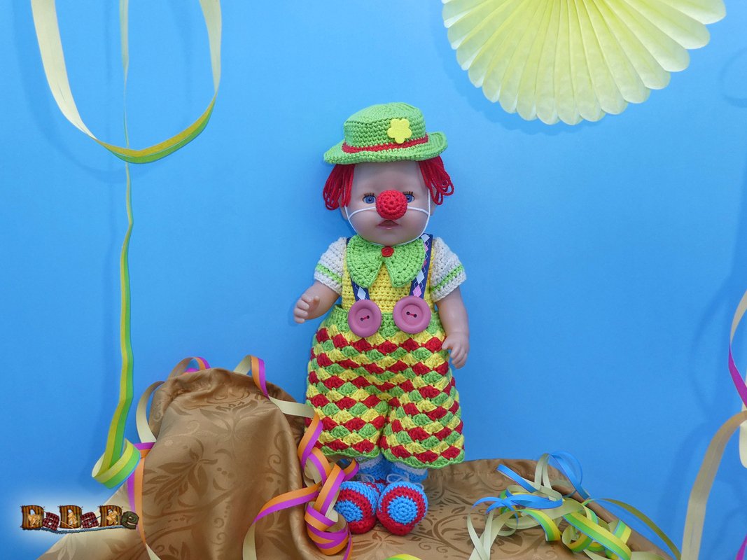 Puppe mit gehäkeltem Clownkostüm: grüner Hut, rote Nase und bunte Latzhose vor blauem Hintergrund