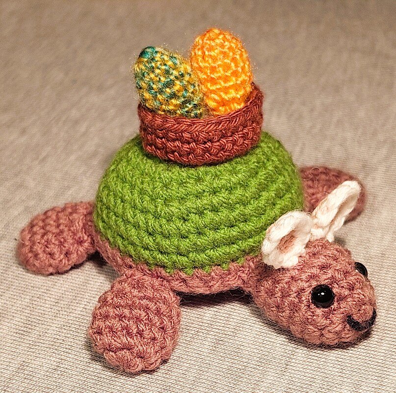 Gehäkelte Amigurumi-Schildkröte mit weißen Hasenohren und Körbchen mit zwei Ostereiern auf dem grünen Panzer