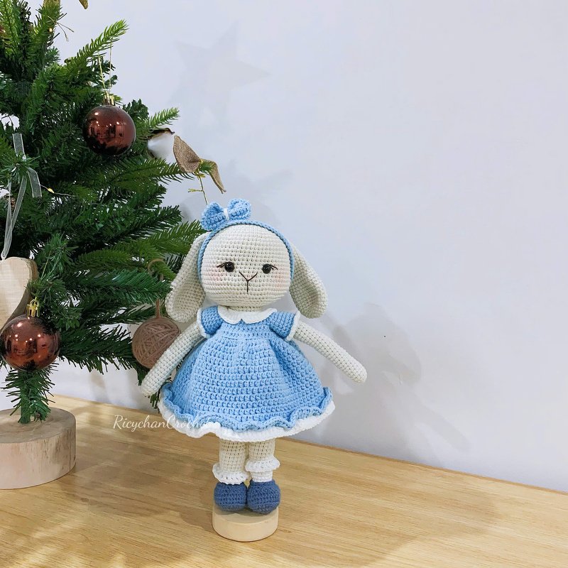 Crocheted amigurumi rabbit in a light-blue dress and headband standing on a wooden base next to a small decorated tree