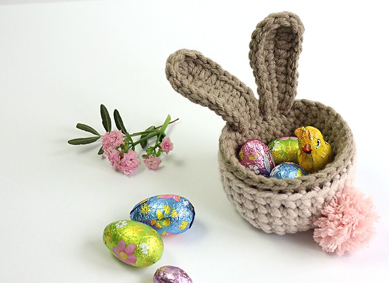 Gehäkeltes Osterkörbchen in Hasenform mit Ohren und rosa Pompom-Schwanz, gefüllt mit bunten Schokoladeneiern