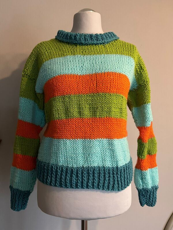 Gestreifter Strickpullover in Grün, Hellblau und Orange mit dunkelblauen Bündchen auf einer Schneiderpuppe.