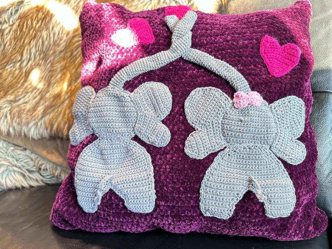 Dunkelviolettes Häkelkissen mit zwei grauen Elefanten, deren Rüssel ein Herz bilden, sowie einem kleinen rosa Herz.