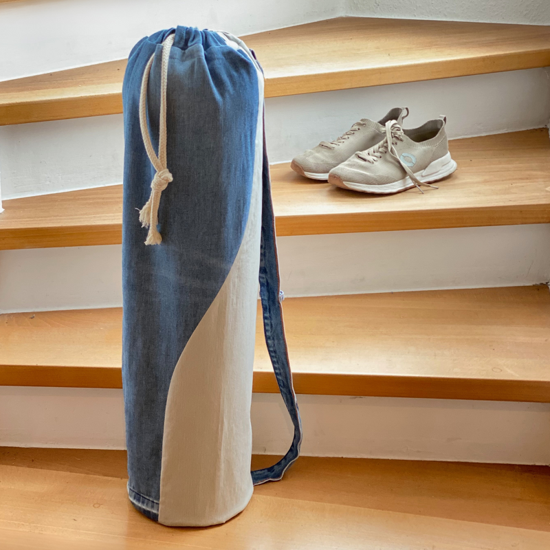 Hohe, schmale Zylindertasche aus blauem Jeansstoff und beigem Stoff mit Kordelzug und Schultergurt auf einer Holztreppe neben beigen Sportschuhen.