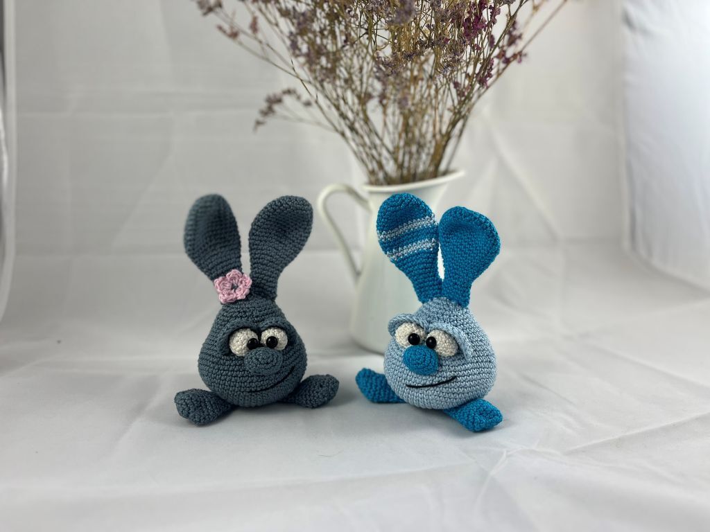 Zwei gehäkelte Amigurumi‑Hasen, links grau mit rosa Blüte am Ohr, rechts blau mit gestreiftem Ohr vor weißer Vase mit Trockenblumen