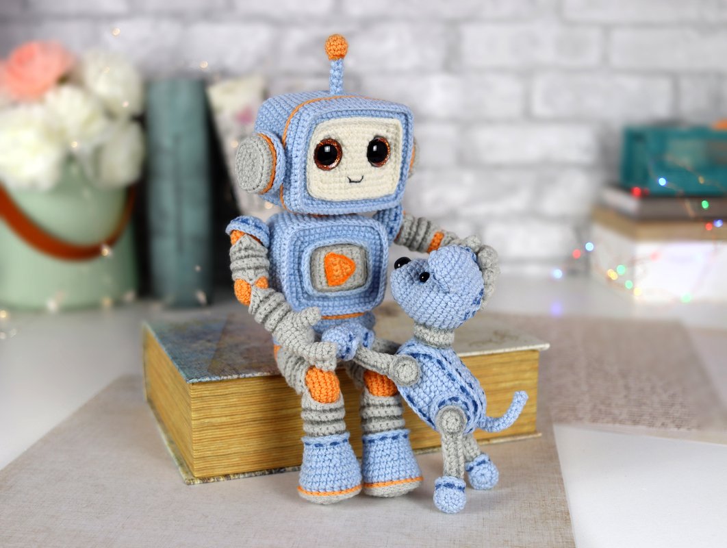 Amigurumi robot and dog crocheted in blue, grey, and orange, sitting on books.
