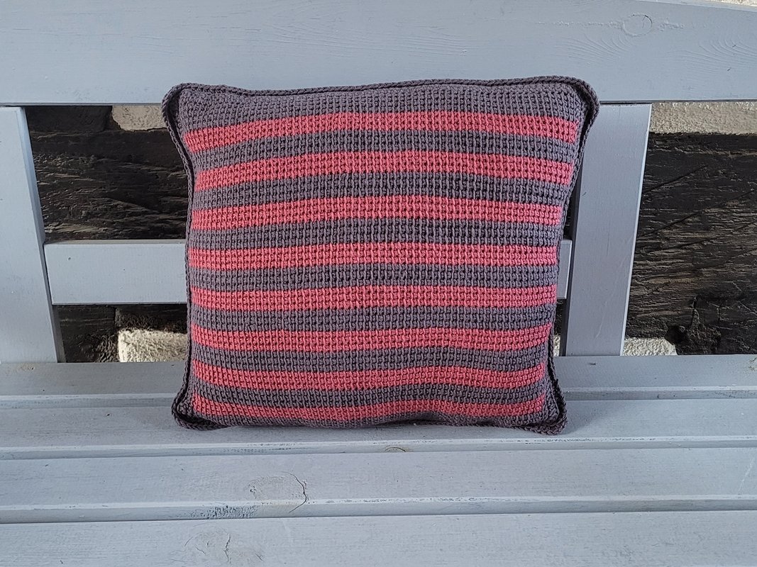 Quadratisches gehäkeltes Kissen mit horizontalen rosa und grauen Streifen auf einer hellgrauen Holzbank