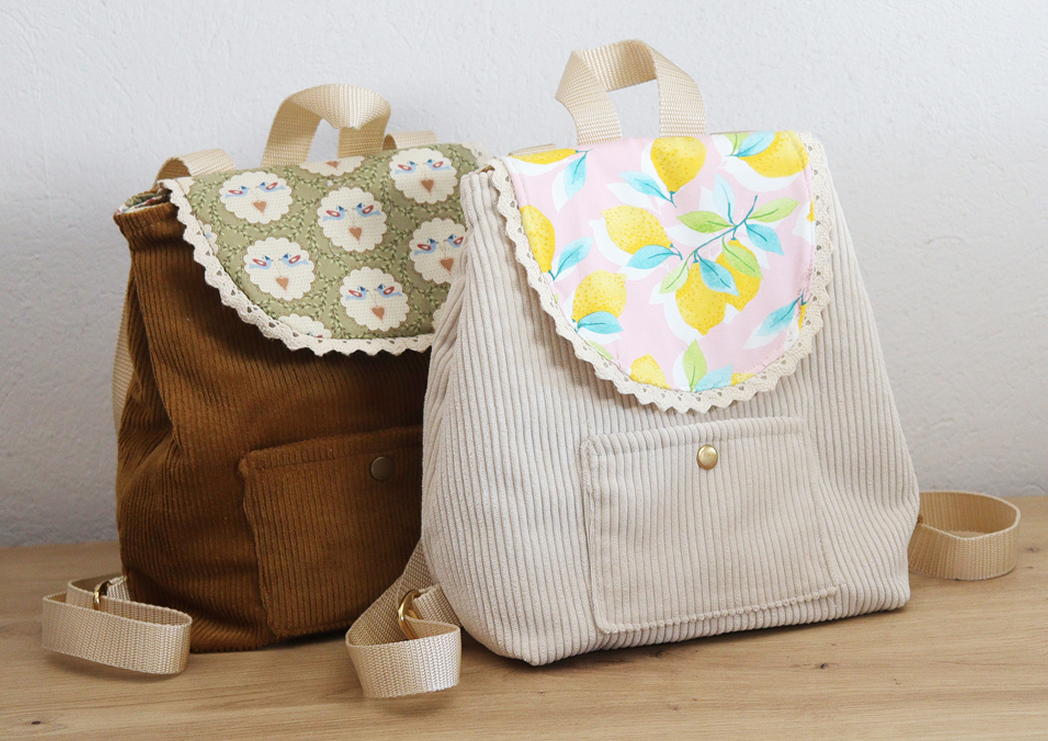 Zwei kleine Kinderrucksäcke aus Cordstoff; vorne hellbeige mit Zitronenprint-Klappe, dahinter braun mit Schäfchenmuster und Spitzenkante