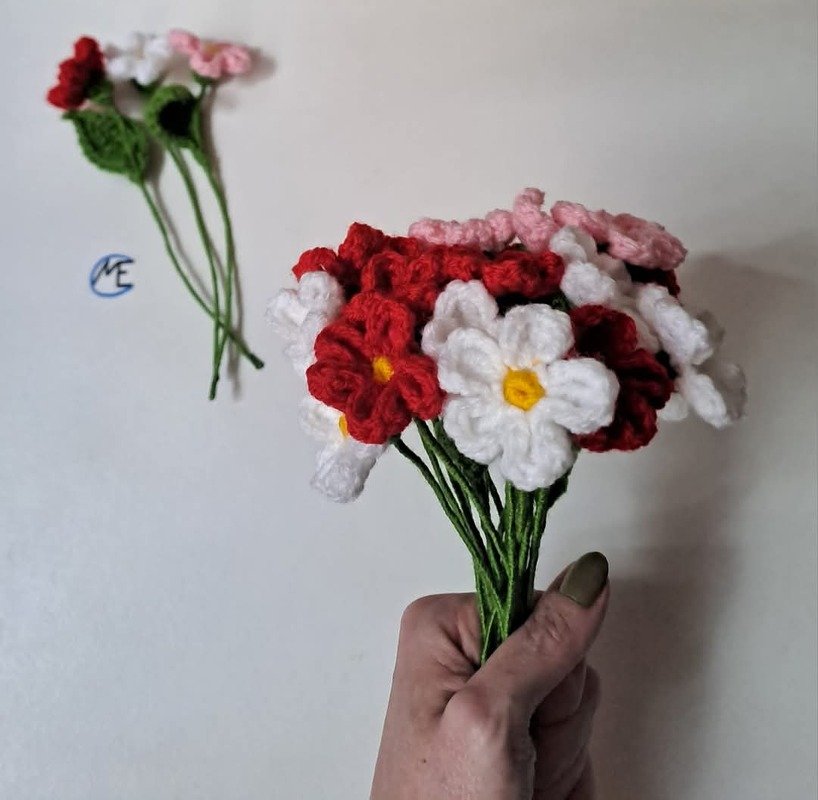 Hand hält handgefertigten Strauß gehäkelter Blüten in Rot, Weiß und Rosa; im Hintergrund liegen weitere Blumen.