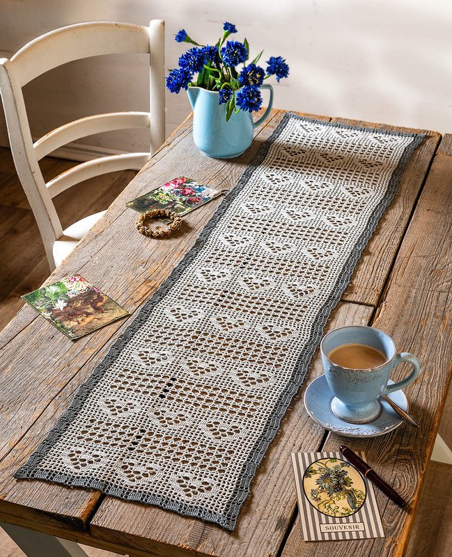 Detailaufnahme eines gehäkelten Tischläufers mit Herz- und Rautenmustern und dunkler Kontrastborte auf einer Holzoberfläche.