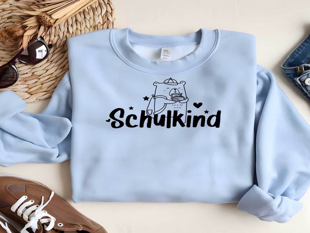 Hellblauer Pullover mit schwarzem Plott: Bär mit Büchern und dem Schriftzug 'Schulkind', umgeben von Sternen und Herz.