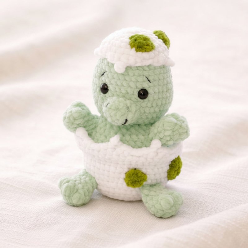 Gehäkelter mintgrüner Amigurumi-Dinosaurier in weißer Eierschale mit grünen Punkten auf hellem Stoff