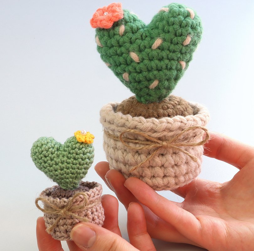 Hands holding two crocheted heart-shaped cacti in small pots with twine bows and tiny flowers