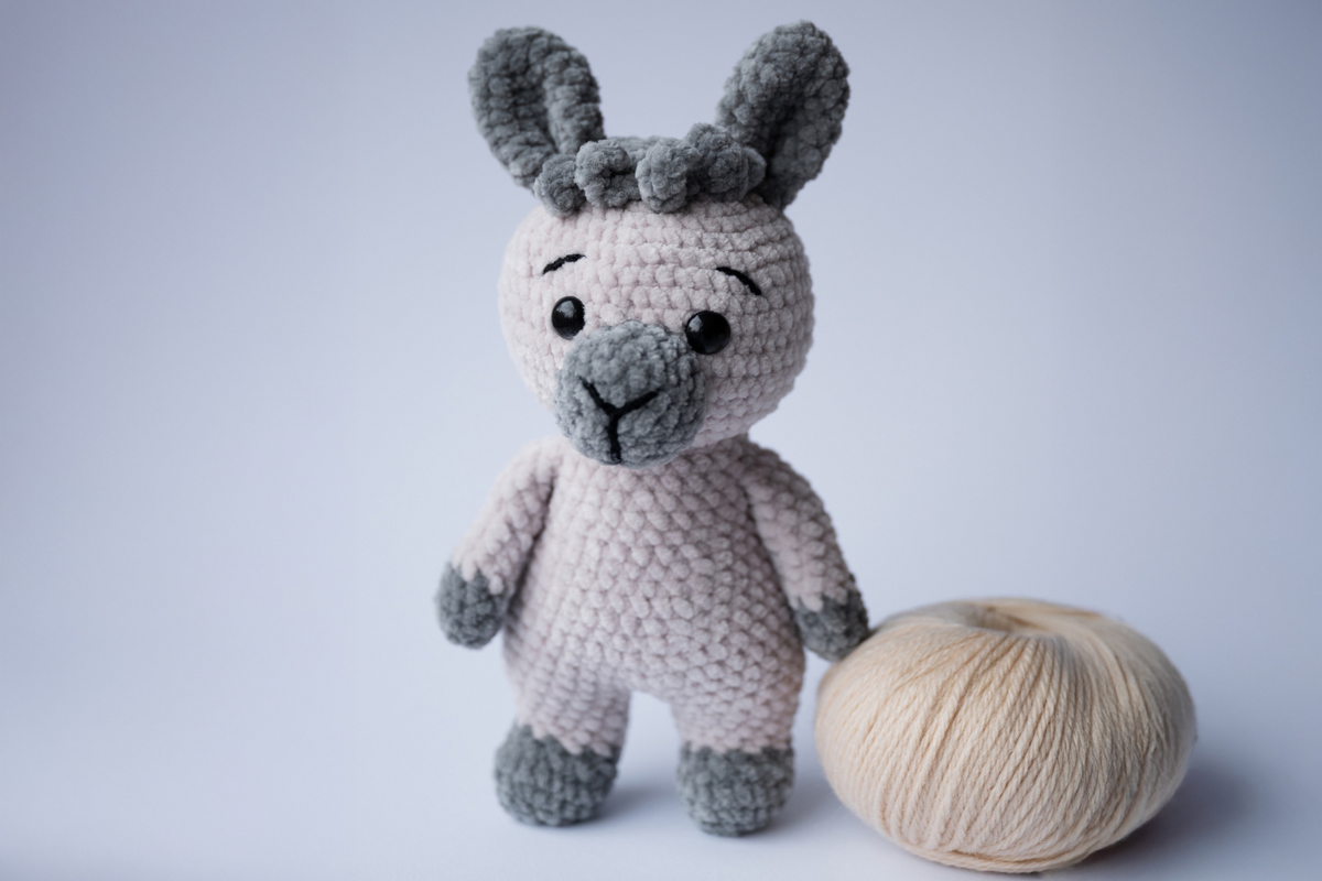 Gehäkeltes Amigurumi-Lama in Rosa und Grau neben einem hellen Wollknäuel auf hellem Hintergrund