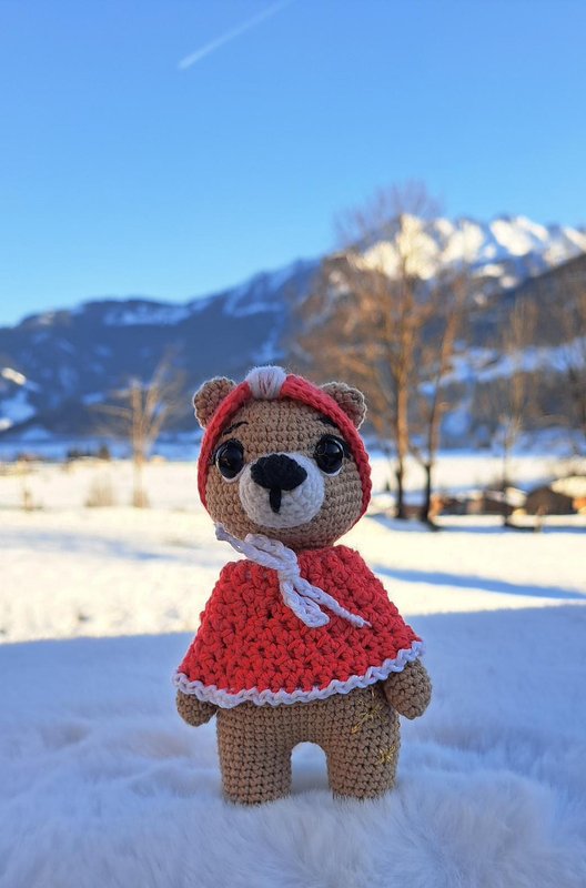 Gehäkelter brauner Amigurumi-Teddybär mit roter Kapuze und weißem Rand vor verschneiten Bergen