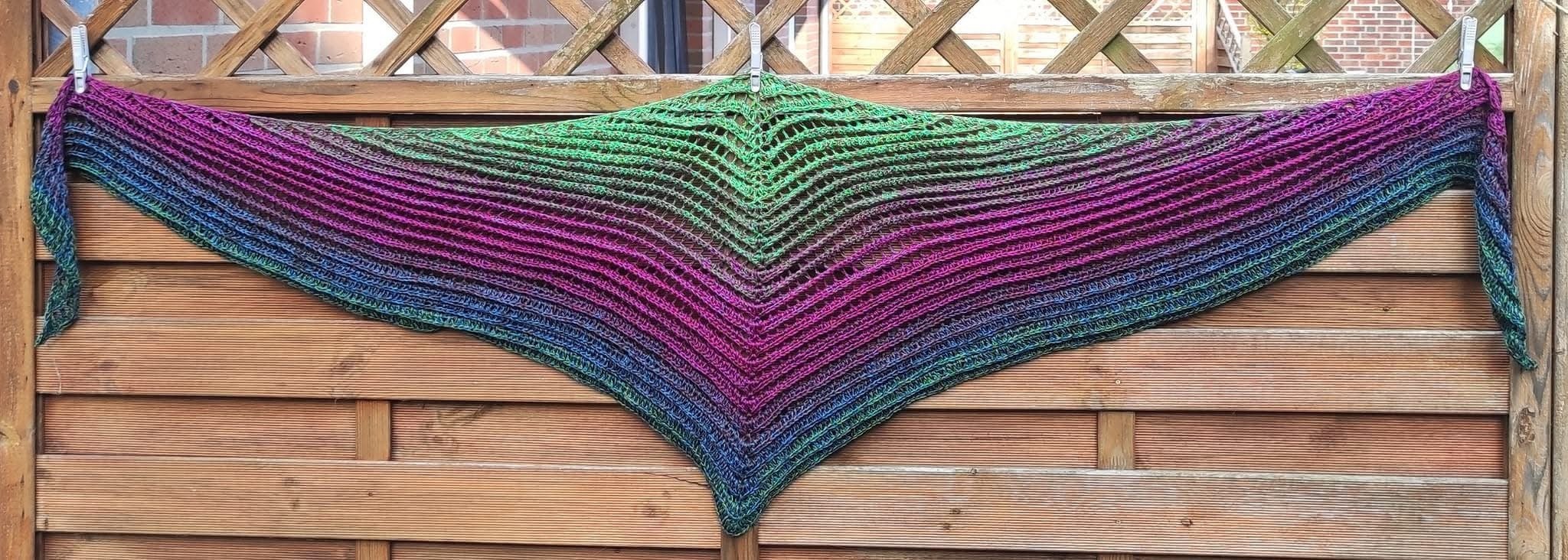 Crochet triangle shawl with green, purple and blue gradient, ribbed texture and openwork top, hung on a wooden fence.