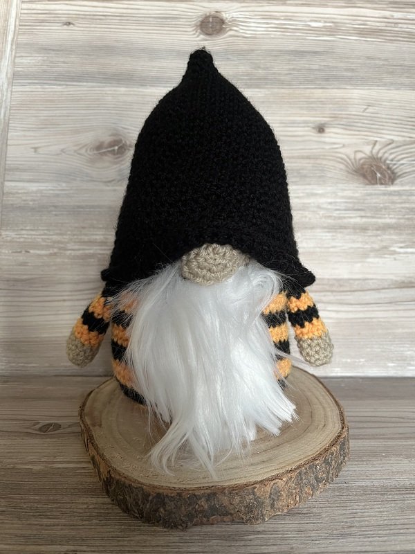 Gehäkelter Wichtel mit schwarzem Spitzhut, weißem Bart und orange‑schwarzen Streifen, stehend auf einer Holzscheibe.