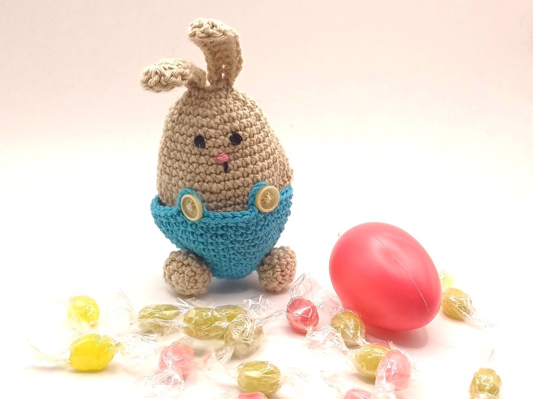 Handgehäkelter beiger Hase in blauer Latzhose, daneben ein rosafarbenes Ei und bunte Süßigkeiten auf hellem Untergrund.