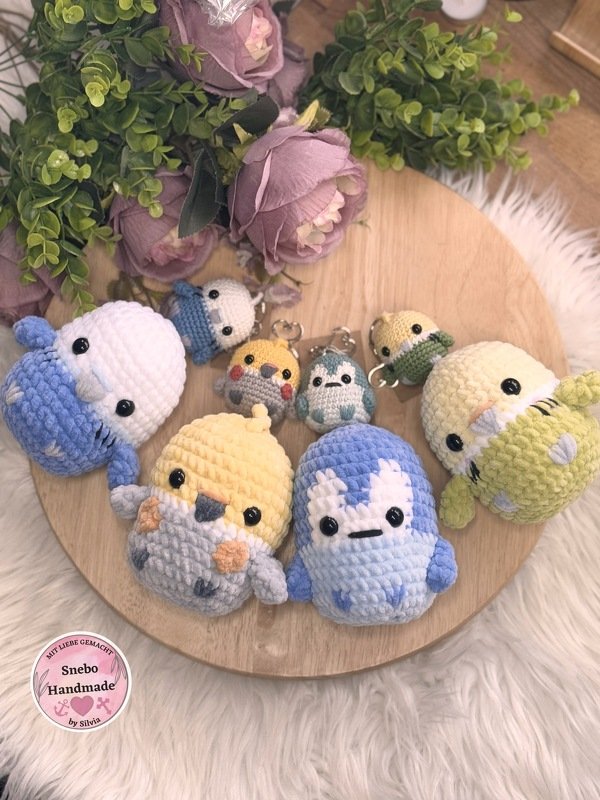 Gehäkelte Amigurumi-Vögel in Pastellfarben, einige als Schlüsselanhänger, arrangiert auf rundem Holzbrett mit Deko