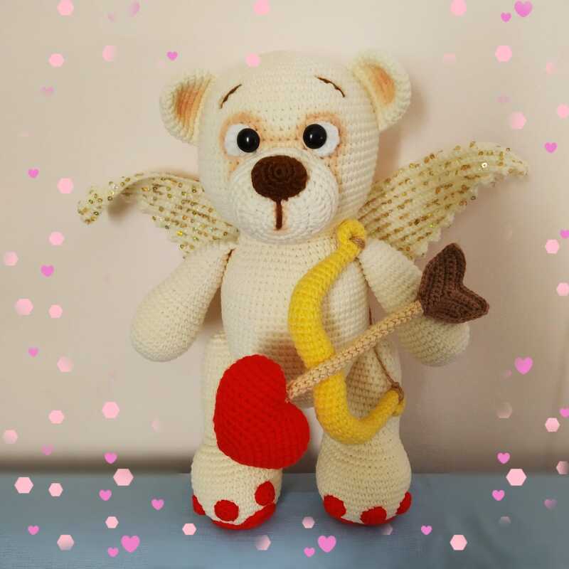 Cream crocheted amigurumi teddy bear with gold-sequined wings holding a yellow bow and a red heart-tipped arrow, red paw pads visible