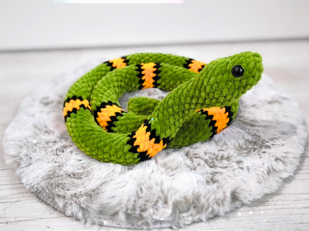 Gehäkelte grüne Amigurumi‑Schlange mit gelb‑schwarzen Streifen auf rundem Fellkissen