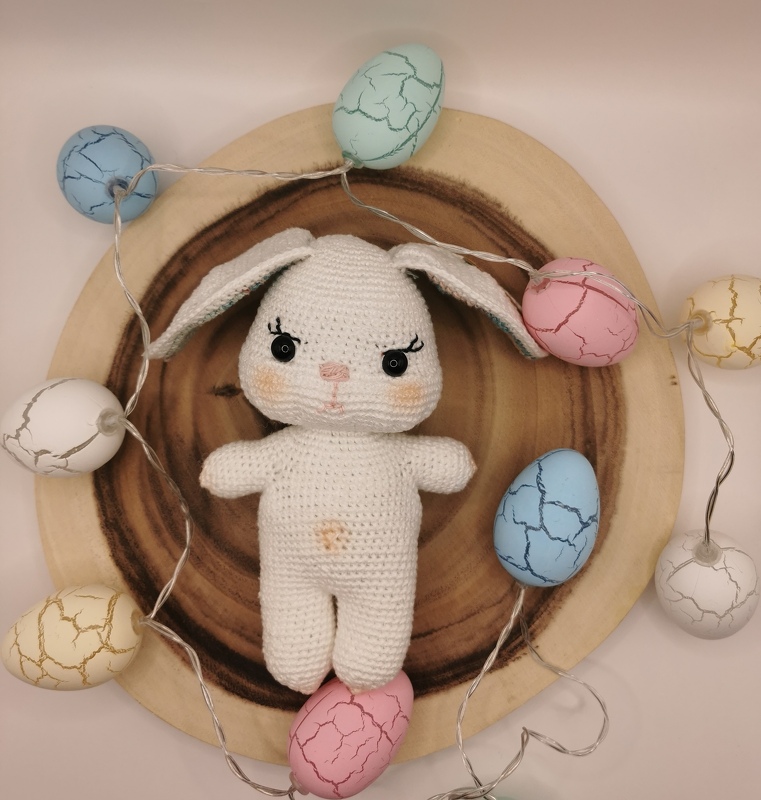 Gehäkelter weißer Amigurumi-Hase auf Holzscheibe, umgeben von pastellfarbenen Ostereiern und Lichterkette