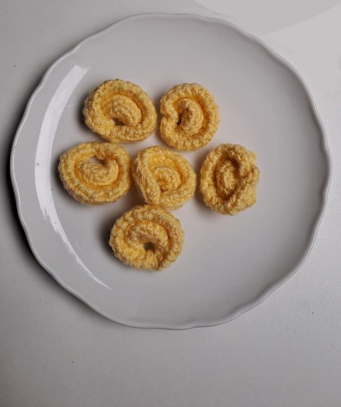 Six small coiled yellow crocheted pieces on a white wavy-edged plate.