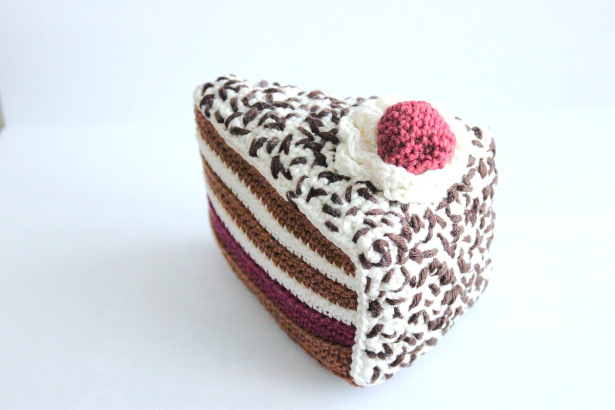 Gehäkeltes Kuchenstück mit weißen und braunen Schichten und rosa Kirsche oben