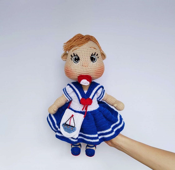 Crocheted amigurumi baby doll with a red pacifier, blue sailor dress, and small bag, held in a hand